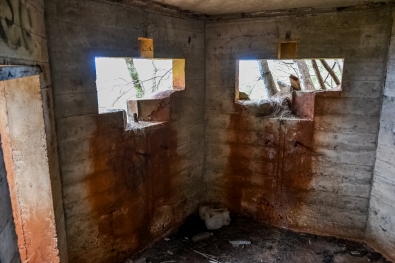 Inside the Bunker. To the left is the open doorway you pass through to gain entrance into it. Ahead of you, cut into the slant-angled forward-facing sides of the bunker, a pair of curious openings. The story behind this architecture is chilling in the extreme. The front faces were slanted to either side, when viewed from the direction of incoming hostile fire, laid down by an amphibious invasion force, as a deflection measure for incoming machine-gun or artillery rounds. The pair of openings are designed to hold the swiveling mounts for 50-caliber machine guns which would be getting used to return fire in an attempt to kill those who were coming ashore below, making their way inland, killing everybody they encountered as they did so. Pear Harbor had been attacked, and World War II was just begun. The fear hovering in the Hawaiian air of the time was thick and palpable, and no one knew what was coming next. Photo by James MacLaren.